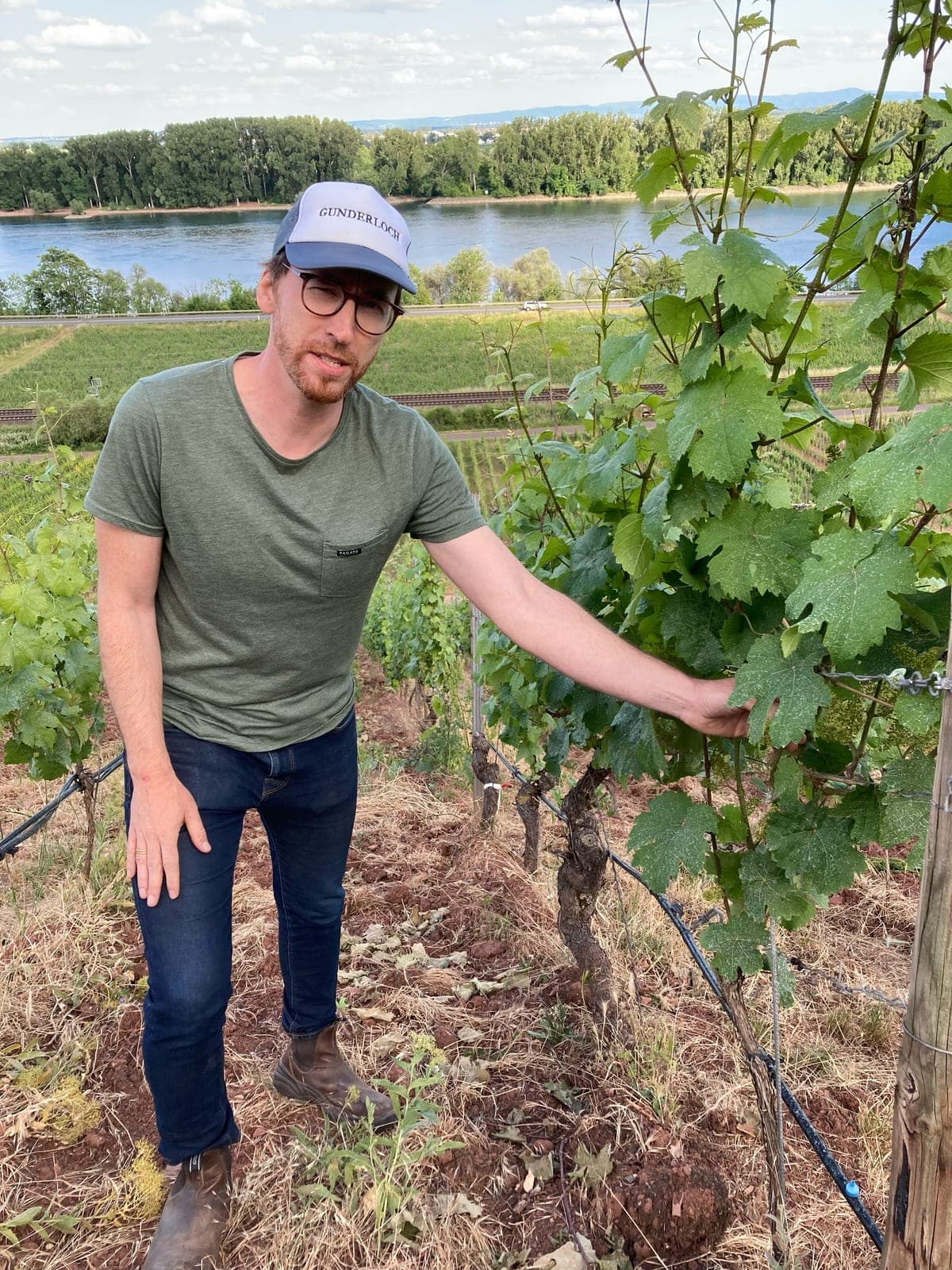 Johannes Hasselbach in a Gunderloch cap and green t-shirt, standing on the red soil of a steep vineyard row with one hand on the vine canopy, the Rhine and the far bank's trees behind him.